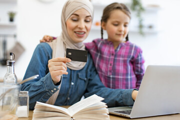 Image depicts family time, online shopping and cooking preparation. Muslim mother and daughter sitting at kitchen table and buying ingredients for meal using laptop, bank card and recipe book.