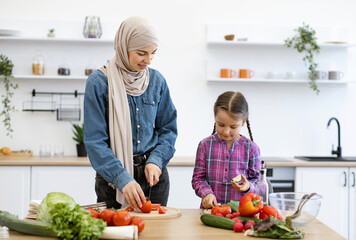 Fresh vegetables on wooden table. Muslim mother and daughter cutting ingredients for salad. Smiling parent teaching child cooking skills spending quality family time together in modern bright kitchen