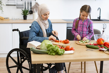 Muslim mother in wheelchair and daughter cutting fresh vegetables for salad in modern kitchen. Enjoying family time, cooking together. Inclusive, love, and nurturing relationship