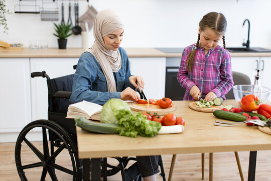 Muslim mother in wheelchair and daughter cutting fresh vegetables for salad in modern kitchen. Enjoying family time, cooking together. Inclusive, love, and nurturing relationship - Powered by Adobe