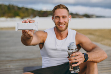 Young strong sporty athletic toned fit sportsman man wear sports clothes eat protein bar drink water warm up training at sunrise sun dawn over sea beach outdoor on pier seaside in summer day morning.