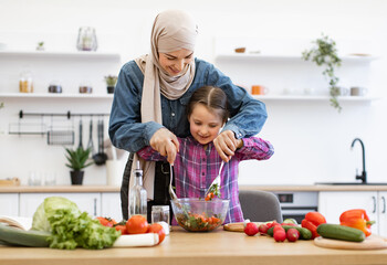Happy and joyful moments of cooking together. Muslim mother and daughter mixing chopped vegetables in salad bowls. Family bonding, healthy eating, homemade meals, and togetherness in modern kitchen.
