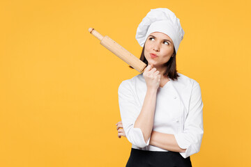 Young minded pensive chef cook baker woman wear white shirt black apron uniform toque chefs hat hold in hand rolling pin look aside on area isolated on plain yellow background. Cooking food concept.