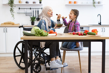 Joyful family bonding moment, healthy lifestyle, and living with disability concept. Muslim mother in wheelchair and daughter eat fresh salad in modern kitchen.