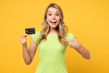 Fototapeta premium Young happy surprised blonde woman she wear green t-shirt casual clothes hold in hand point finger on mock up of credit bank card isolated on plain yellow orange background studio. Lifestyle concept.