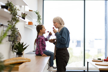 Muslim mother and daughter enjoying healthy salad together in modern kitchen, showcasing family bonding and healthy lifestyle. Mother wears hijab, daughter smiles while eating.