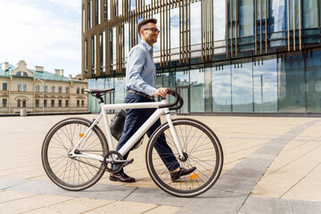 A man in business attire walks his bicycle past a modern office building, embracing a sustainable...