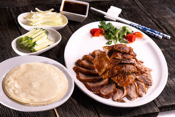 Peking Duck on a white plate served with fresh cucumber, green onions, cilantro and roasted wheaten chinese pancakes on black  wooden background. Chinese food. food concept.
