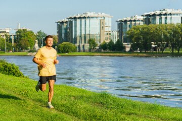 Fototapeta premium A man in a yellow shirt jogs along a lush riverside path, enjoying the greenery and water views in an urban park.