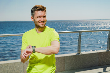 Man in a bright green sports shirt checks his smartwatch while jogging along a sunny seaside...