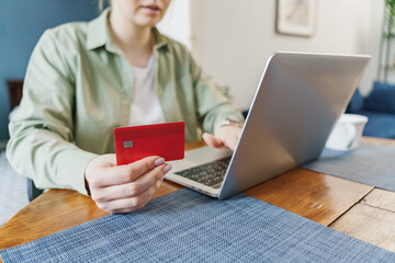 A person holds a red credit card while using a laptop, focused on online shopping or banking in a home setting.