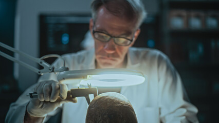 Senior archaeologist takes measurements of fossil skull using professional measuring tool, looks through magnifying lamp at the remains of extinct prehistoric human in archaeological lab at night.