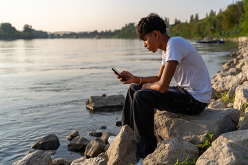 portrait of young teenage man sitting on river bank at sunset using smart phone