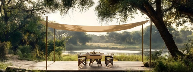 A simple canopy with two thin wooden poles and beige fabric hanging over an outdoor dining table, surrounded by trees overlooking an African river in a safari-style setting.