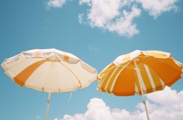 Two beach umbrellas in vibrant orange and white colors against a bright blue sky with scattered clouds Perfect for summer themes and vacation imagery