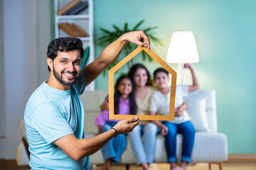 Indian dad holding a cardboard home cutout as his family sits on the sofa, real estate dreams