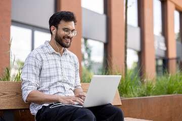 A smiling young Muslim man sits on a bench outside in the street wearing headphones, working on a...
