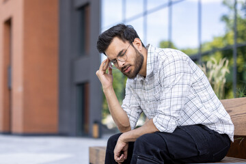 Obraz premium Tired young Indian man sitting on a bench outside an office building and holding his head with his hand, feeling a lot of pain and stress