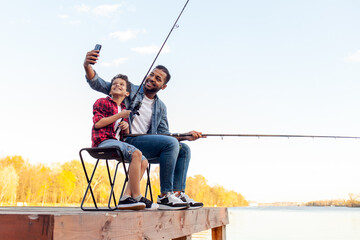 An African American father and son sit on a wooden pier by a lake enjoying a fishing trip. The...