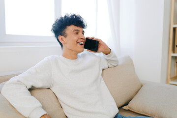 young man talking on smartphone, sitting on a cozy sofa in a bright, modern room with natural light streaming in through the window Relaxed atmosphere with a cheerful expression