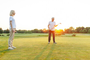Obraz premium old senior couple in white clothes playing golf on golf course and chatting at sunset, elderly man and woman actively relaxing outdoors and laughing, pensioners on vacation
