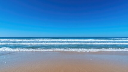 Obraz premium Clear blue sky over a pristine sandy beach with gentle waves lapping at the shore, evoking a classic summer day