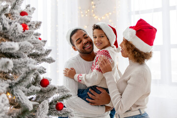 African American family in Santa hat celebrate New Year together at home and decorate Christmas tree, teenage boy celebrates New Year with mom and dad and holds red toy