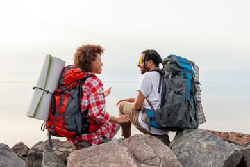 young african american couple tourists with hiking equipment and backpacks sitting on top of...