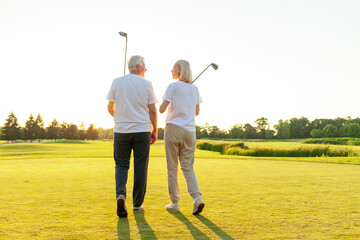elderly senior couple in uniform holding golf clubs and walking on the background of golf course at sunset and talking, old man and woman walking and talking on golf game and looking at copy space