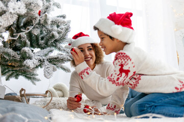 african american teenage boy in santa hat decorates christmas tree at home with mom, child celebrates new year and having fun with parent at christmas