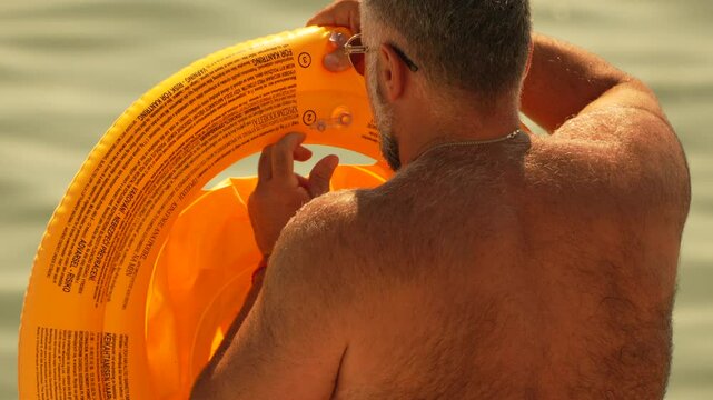 Man Inflates Floatation Device Beach Summer. A man inflates a yellow inflatable ring in shallow water. Slow motion