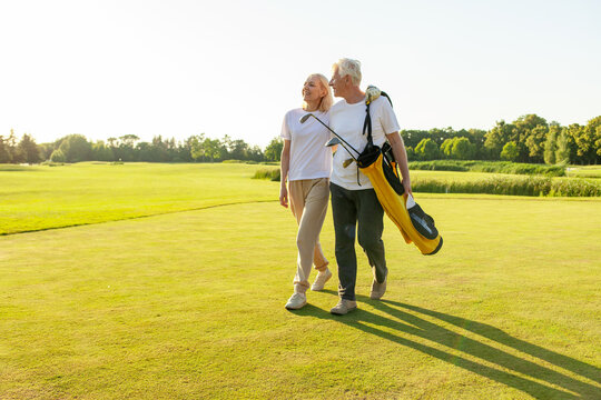 elderly senior couple in uniform holding golf clubs and walking on the background of golf course at sunset and talking, old man and woman walking and talking on golf game and looking at copy space