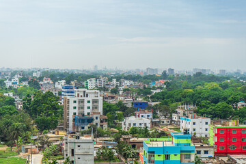 Obraz premium cityscape with multiple buildings of varying heights and colors, interspersed with dense vegetation under a hazy sky. Rajshahi, Bangladesh