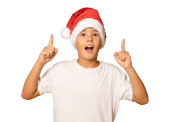 Portrait of a surprised boy in a red hat and white t-shirt pointing fingers up on a transparent background. A child in a Christmas hat of Santa Claus.