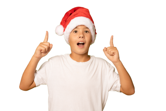 Portrait of a surprised boy in a red hat and white t-shirt pointing fingers up on a transparent background. A child in a Christmas hat of Santa Claus. - Powered by Adobe
