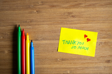 Vista superior de nota adhesiva amarilla con agradecimiento y corazones junto a rotuladores de colores con fondo de madera