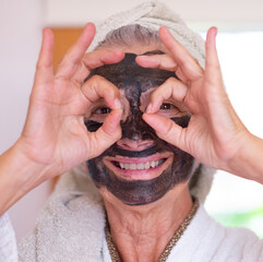 Obraz premium Joyful cheerful senior woman with a detox facial charcoal mask homemade and a towel on head smiles looking at camera through her fingers - take care of the skin concept