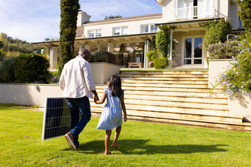 Carrying solar panel, father and daughter walking towards house on sunny day