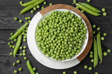 Fresh green peas in bowl and pods on black wooden table, flat lay