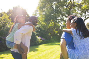 Fototapeta premium Carrying daughters piggyback, multiracial parents smiling and enjoying family time in sunny park