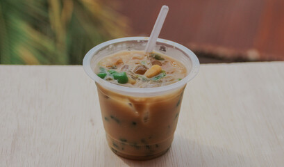 A cup of traditional indonesian iced drink green cendol with brown sugar and coconut milk with a spoon on a wooden table
