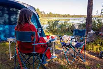 Woman, sitting on a camping chair in front of a tent, enjoying cup of coffee and amazing view in...