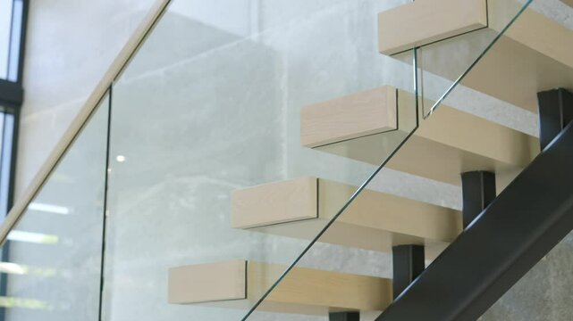 Transparent glass railing with wooden handrail for freshly built staircase by wall with concrete tiles. Minimalistic steps leading on floor down in grey space