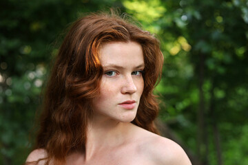 Naklejka premium Portrait of ginger girl with long red hair and freckles in park. Young sensual model with naked shoulders, natural female beauty