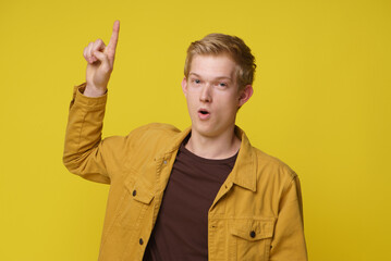 A young man in a casual yellow jacket smiles and points his index finger to the side, against a bright yellow backdrop