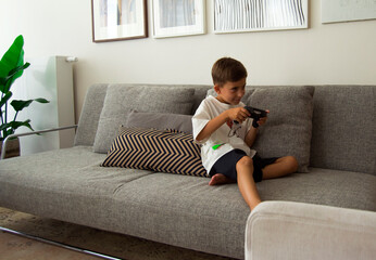 Cute 8 year old boy playing video game console while sitting on couch at home.