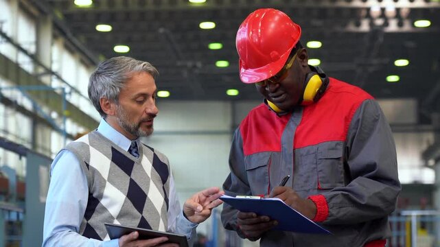 Supervisor Signing a Document in a Factory and Talking to Black Worker in Protective Workwear - Zoom Out. Caucasian Production Manager Giving Instructions to Black Industrial Worker.