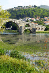 View from Trebinje, Bosnia and Herzegovina: grass on the bank, waters of the Trebišnjica River, arches of the Arslanagic stone bridge and Crkvice peak with the Hercegovacka Gračanica monastery.