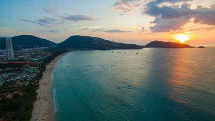 A breathtaking aerial view of a coastal bay at sunset, with golden light reflecting on the calm sea and silhouetted hills in the distance. beautiful reflection of the golden sun at Patong Bay Phuket.