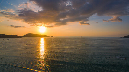 A breathtaking aerial view of a coastal bay at sunset, with golden light reflecting on the calm sea and silhouetted hills in the distance. beautiful reflection of the golden sun at Patong Bay Phuket.